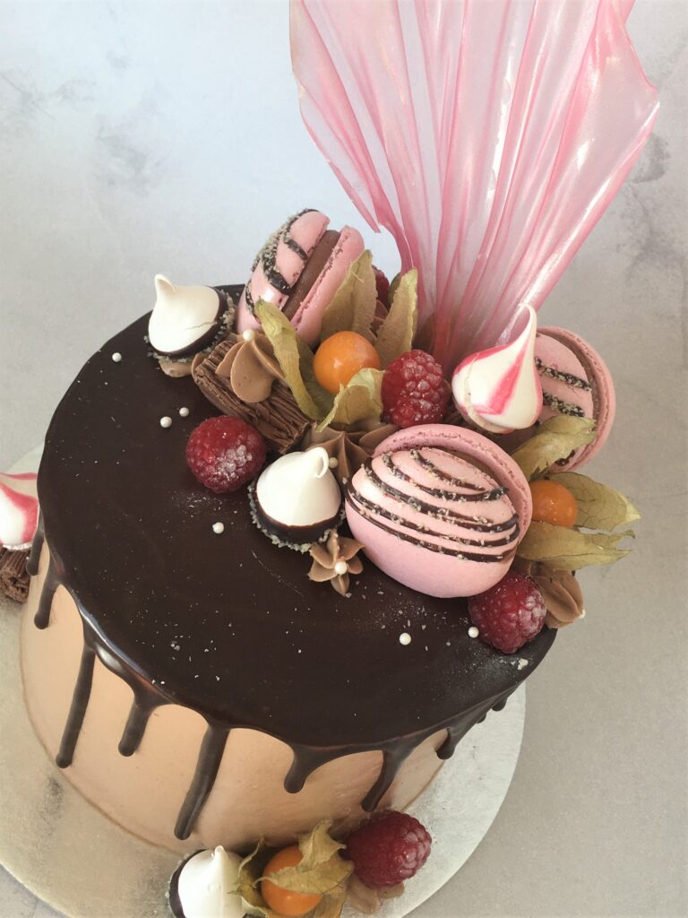 Chocolate drip cake decorated with macarons, meringue kisses, flake, fresh raspberries and rice paper sail